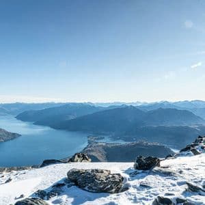 Cima di montagna innevata che domina un grande lago serpeggiante e catene montuose lontane sotto un cielo luminoso e soleggiato.
