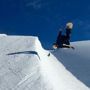 Uno snowboarder con casco esegue una acrobazia a testa in giù a mezz'aria sopra un halfpipe di neve sotto un cielo blu sereno.