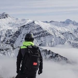 Una persona con uno zaino e un casco in cima a una montagna che ammira le cime innevate sopra un mare di nuvole