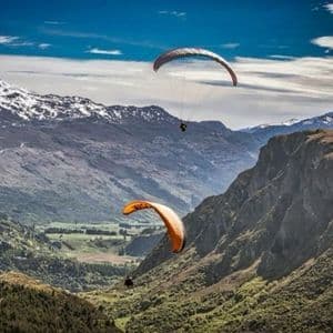 Due parapendii che sorvolano una valle verde circondata da montagne aspre con cime innevate sotto un cielo azzurro.