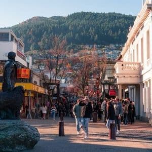 Persone che camminano lungo una strada pedonale fiancheggiata da negozi e da una statua, con una collina boscosa sullo sfondo.
