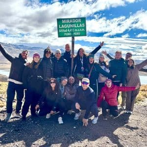 Un viaggio di gruppo WeRoad in posa all'aperto accanto a un cartello alpino verde con montagne e un lago alle loro spalle