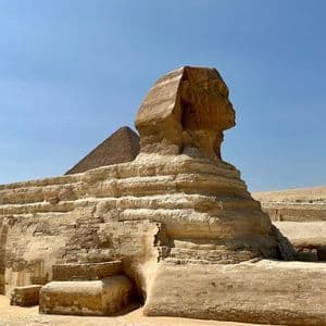 Seitenprofil eines massiven Steinsphingenmonuments in einer Wüstenlandschaft mit einer Pyramide im Hintergrund