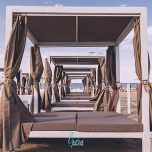 Una fila di cabine da spiaggia in legno bianco con tende e lettini imbottiti su una riva sabbiosa sotto un cielo luminoso