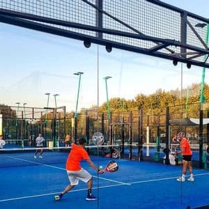 Persone che giocano a padel su un campo blu all'aperto racchiuso da pareti di vetro e recinzione metallica