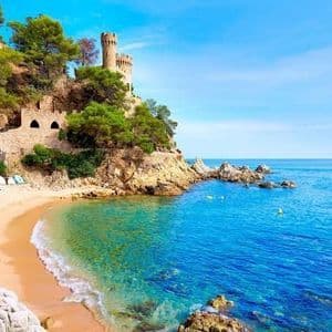 Un castello di pietra sorge su una scogliera rocciosa e alberata che domina una caletta sabbiosa con acqua di mare turchese sotto un cielo limpido