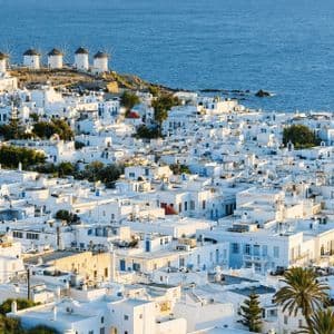 Edifici di una cittadina costiera imbiancati a calce si estendono su una collina vicino al mare blu con i mulini a vento sullo sfondo.