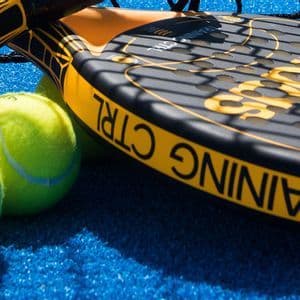 Due racchette da padel nere e gialle e palline verde brillante riposano su un campo in erba sintetica blu sotto l'ombra della rete