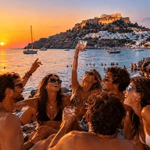 Un viaggio di gruppo WeRoad con drink su una spiaggia rocciosa al tramonto, con barche e un borgo collinare costiero sullo sfondo.