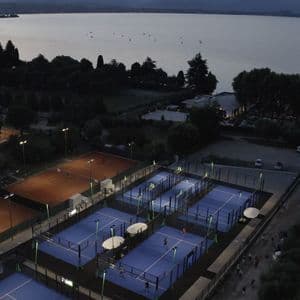 Illuminated blue padel courts and clay tennis courts beside a large lake at dusk, viewed from above.