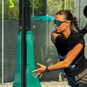 A woman wearing sunglasses prepares to hit a padel ball with her racket on an outdoor court.