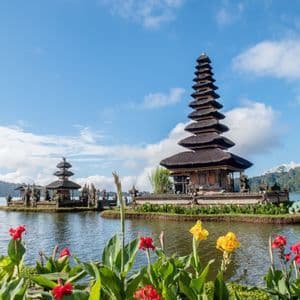 Deux temples traditionnels aux toits étagés se dressent sur un lac, avec des fleurs rouges et jaunes au premier plan sous un ciel partiellement nuageux.