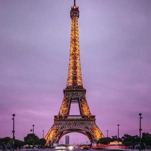 La Torre Eiffel, iluminada con luces doradas, se ve desde una calle con estelas de luces de tráfico bajo un cielo púrpura al anochecer.