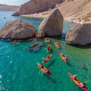 Un viaggio di gruppo WeRoad in kayak su canoe rosse attraverso limpide acque turchesi vicino a grandi massi e una spiaggia.