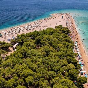 Vista aerea di una lingua di spiaggia triangolare fiancheggiata da pini e bagnanti circondata da acque limpide e blu.