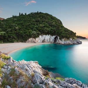 Una cala de arena con aguas tranquilas y turquesas, resguardada por un acantilado boscoso con una cueva marina al atardecer.