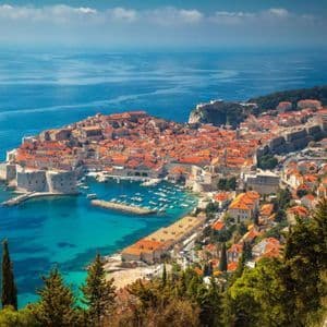 Una vista panoramica di una città costiera murata con tetti in terracotta, circondata da un mare blu calmo e un porto pieno di barche.