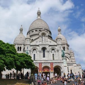 Folla di persone siede e cammina sui gradini all'esterno di una grande basilica con cupola bianca sotto un cielo parzialmente nuvoloso.