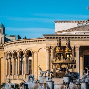 Una fuente de piedra con estatuas de animales y un caballo dorado se encuentra frente a un gran edificio con columnatas clásicas.