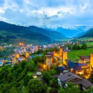 Torres y casas de piedra medievales iluminadas dispersas por un valle montañoso verde y frondoso al atardecer.
