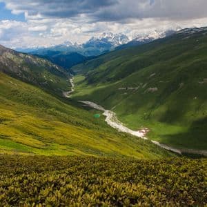 Un valle montana verde con un río serpenteante que se dirige hacia picos nevados bajo un cielo parcialmente nublado.