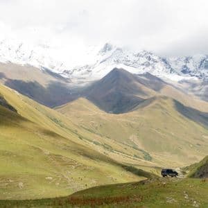 Una furgoneta oscura aparcada en una colina cubierta de hierba con vistas a un amplio valle y picos de montañas nevadas al fondo.