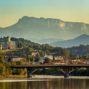Un puente arqueado cruza un río tranquilo que conduce a un pueblo con una catedral en la colina frente a grandes montañas.