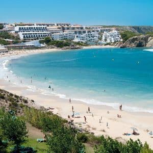 Une large vue d'une crique de sable où l'on se baigne dans des eaux turquoise, avec des bâtiments blancs sur les falaises au-dessus de la plage.