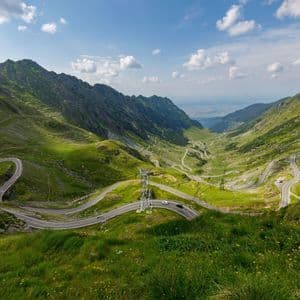 A winding road with cars travels through a lush green mountain valley under a blue sky with some clouds.