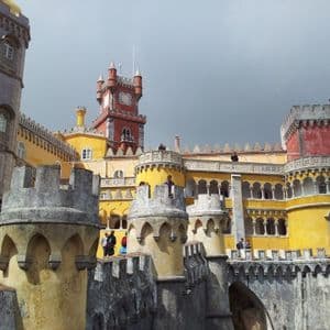 A colorful castle with yellow and red towers, viewed from its stone ramparts where people are walking under a cloudy sky.