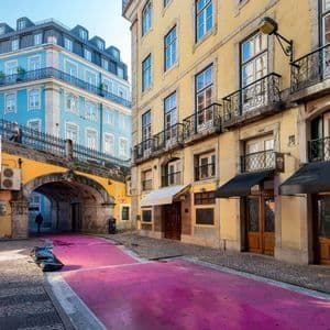 Una stretta via di città con una strada rosa brillante che passa sotto un arco in pietra, tra edifici gialli e blu.