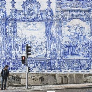 Una persona è in piedi su un marciapiede, accanto a un semaforo, davanti a un grande muro ricoperto di intricate piastrelle blu e bianche.