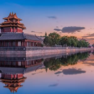 A traditional Chinese corner tower on a stone wall is reflected in the calm water of a moat at sunset.