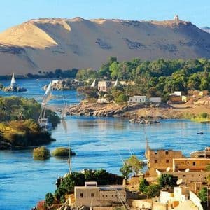 Un amplio río azul con veleros, bordeado por un pueblo con palmeras y una gran duna de arena dorada.