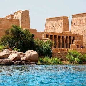 Un antico tempio di pietra con pilastri e colonne intagliate si erge su una sponda rocciosa e verdeggiante accanto ad acque di un blu brillante.