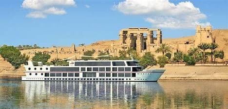 Una nave da crociera bianca galleggia su un fiume di fronte a rovine antiche e palme su una riva sabbiosa sotto un cielo blu.