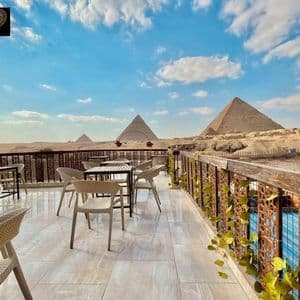 A cafe terrace with empty tables and chairs overlooks the great pyramids in a desert landscape under a blue sky with clouds.