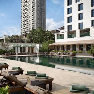 Tumbonas vacías con toallas verdes enrolladas están alineadas junto a una piscina larga con edificios modernos al fondo.