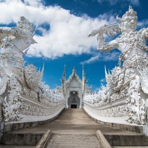 Vue en contre-plongée d'une allée menant à un temple blanc orné, encadré de grandes statues complexes sous un ciel bleu.