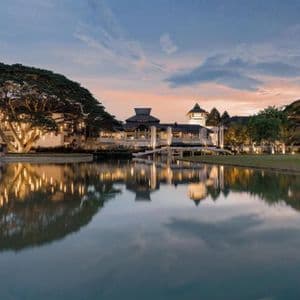 Un complexe hôtelier illuminé à l'architecture traditionnelle se reflète dans un lac calme au crépuscule, à côté d'un grand arbre décoré.