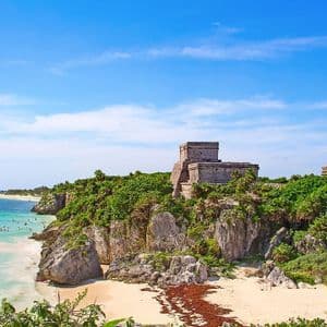 Antiche rovine in pietra si ergono su una scogliera verdeggiante, affacciate su una spiaggia sabbiosa e un mare turchese sotto un cielo azzurro.