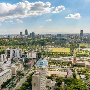 Luftaufnahme einer weitläufigen Stadtlandschaft mit modernen Wolkenkratzern und üppigen grünen Parks unter einem teilweise bewölkten blauen Himmel.