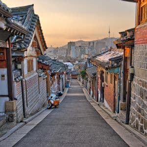 A narrow street slopes downhill between traditional houses with tiled roofs, revealing a modern city skyline in the distance at sunset.