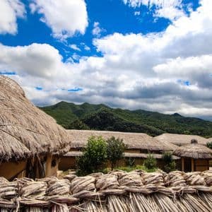 Un villaggio di capanne tradizionali dai tetti di paglia si trova ai piedi di montagne verdi, sotto un cielo parzialmente nuvoloso.