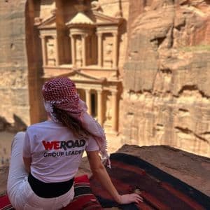 Ein WeRoad Gruppenleiter mit einer Keffiyeh sitzt auf einem Felsvorsprung mit Blick auf ein antikes Gebäude, das in eine rote Felswand gehauen ist.