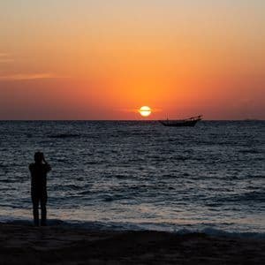 Une personne en silhouette sur une plage photographie le coucher de soleil orange sur l'océan, avec un bateau à l'horizon.