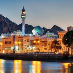 Une mosquée illuminée avec un dôme en mosaïque bleue et un minaret sur un front de mer, avec des montagnes derrière la ville au crépuscule.