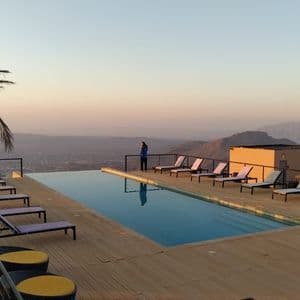 Una mujer de pie junto a una piscina infinita en una terraza de madera, contemplando un valle montañoso al atardecer.