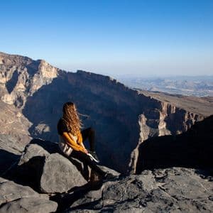 Une femme aux cheveux longs est assise sur un rebord rocheux, dominant un vaste canyon ensoleillé sous un ciel bleu clair.
