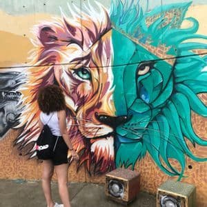 Une femme avec un sac à dos WeRoad contemple une fresque de street art colorée, un visage de lion, sur un mur en béton.